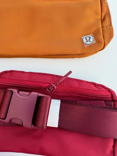 بلت بگ Lululemon، نمای نزدیک سگک و زیپ کیف زرشکی، کنار کیف نارنجی با نشان لولولمون