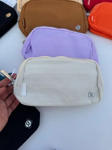 بلت بگ Lululemon، خاکستری بسیار روشن با نشان فلزی لولولمون؛ در دست کنار کیف‌های رنگی