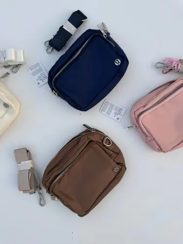 کیف ساید سینچ Lululemon، کیف‌های سرمه‌ای، قهوه‌ای و صورتی با لوگوی فلزی و بندهای جدا روی سطح سفید