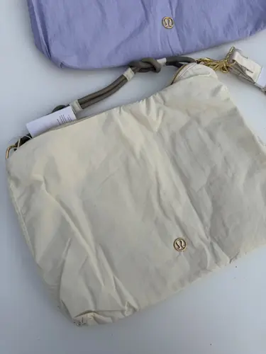 کراس بادی کانورتیبل Lululemon، کیف کرم با لوگوی طلایی و دسته خاکستری کنار کیف بنفش روشن روی سطح سفید