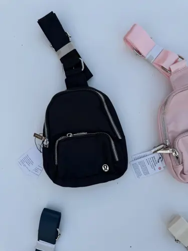 کیف اسمال اسلینگ Lululemon، مشکی با جیب زیپ‌دار جلو و بند پهن، کنار کیف صورتی