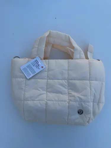 کیف مینی توت بگ Lululemon، کیف مینی کرم با دوخت مربعی، دسته‌های کوتاه و لوگوی فلزی روی سطح آبی روشن