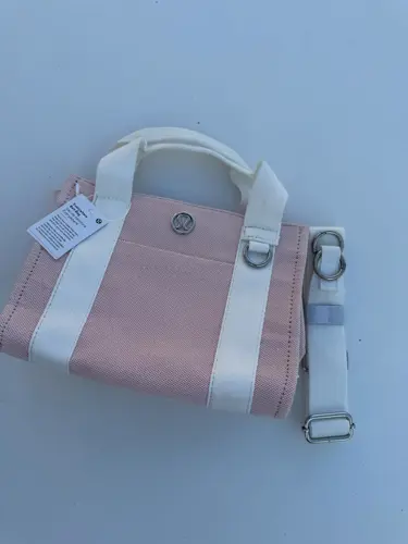 کیف کانواس Lululemon، صورتی روشن با دسته و نوارهای سفید، نشان فلزی لولولمون و بند جداشونده، روی زمینه روشن