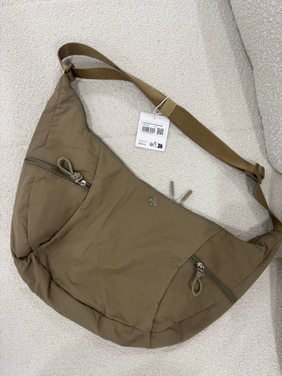 کیف اسلوچی اسلینگ 13 لیتری Lululemon، کیف اسلینگ زیتونی با جیب بیرونی و بند بلند روی سطح روشن