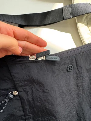 کیف اسلوچی اسلینگ Lululemon 13L مشکی؛ نمای زیپ دهانه، لوگو و قطعه تنظیم بند در دست