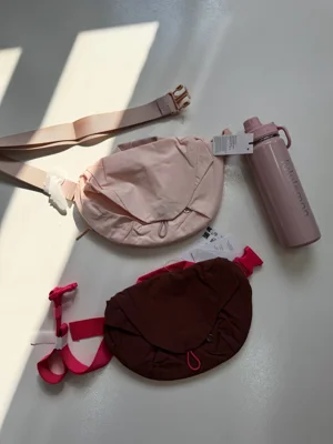کراس بادی کرو Lululemon صورتی با بند پهن و بطری صورتی کنار آن؛ کیف قهوه‌ای در پایین تصویر - تصویر اصلی
