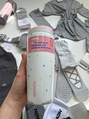 کافی کاپ Owala 20oz سفید با ستاره‌های رنگی، لوگوی صورتی و برچسب ظرفیت ۵۹۰ میلی‌لیتر در دست
