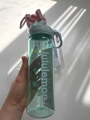 باتل سبز شفاف Lululemon با نوشته روشن، درب نی‌دار و حلقه حمل در دست، کنار مدل آبی و صورتی