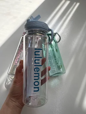 باتل شفاف Lululemon با نوشته آبی، درب نی‌دار تاشو و حلقه حمل در دست، کنار مدل‌های صورتی و سبز