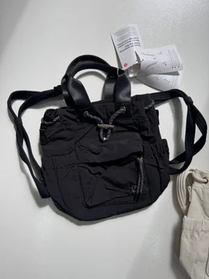باکت بگ کانورتیبل مینی Lululemon 5L مشکی؛ نمای واقعی جلو و بندهای حمل، با بخشی از کیف شیری کنار آن