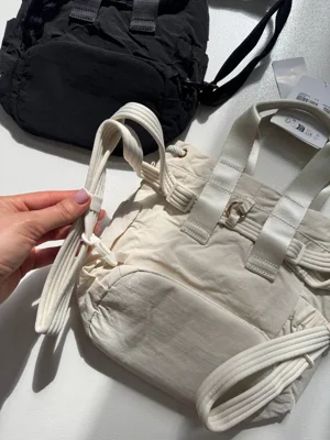 باکت بگ کانورتیبل مینی Lululemon 5L شیری؛ نمای مایل جلو و جیب کناری با بند حمل، در کنار کیف مشکی