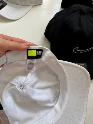 لیبل HERITAGE86 و ONE SIZE FITS MOST داخل کلاه Nike سفید؛ کلاه مشکی در پس‌زمینه