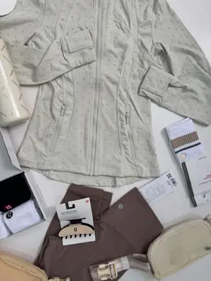 دیفاین جکت لون Lululemon، جکت خاکستری روشن طرح‌دار زیپ‌دار، پهن‌شده کنار کلاه، بطری، جوراب‌ها و کیف‌های روشن