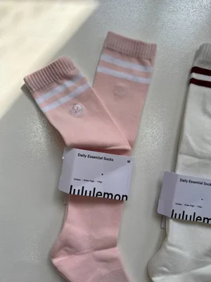 نمای نزدیک جوراب Lululemon Knee-High صورتی با دو خط سفید، لوگوی دوخت‌شده و برچسب سایز M