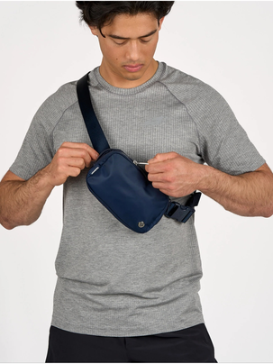 بلت بگ وردمارک Lululemon، کیف کمری سرمه‌ای به‌صورت مورب روی مدل با تیشرت خاکستری، در حال بازکردن زیپ
