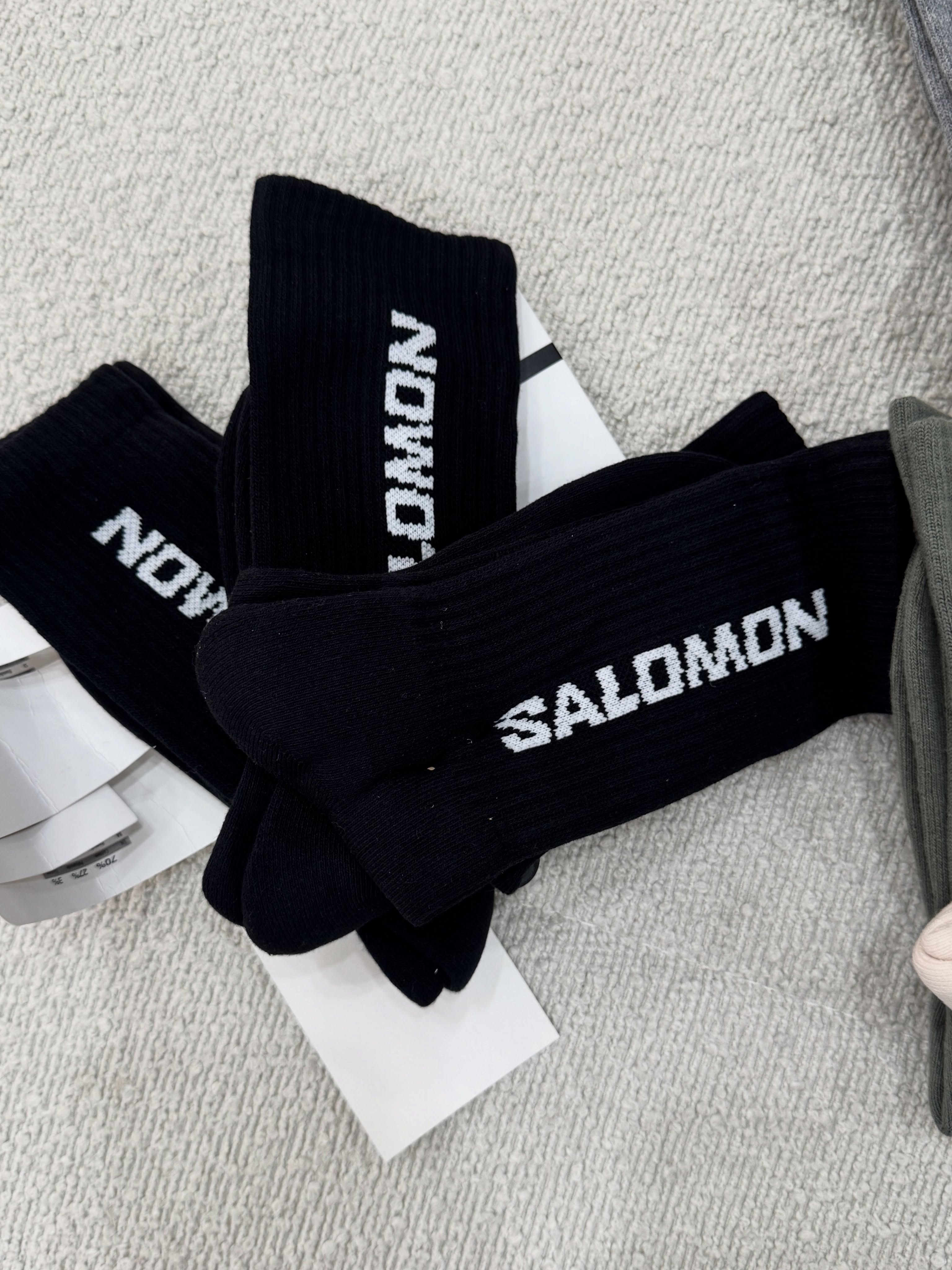 پک سه‌جفتی جوراب ساق بلند مشکی Salomon با نوشته سفید و بافت کبریتی روی زمینه روشن