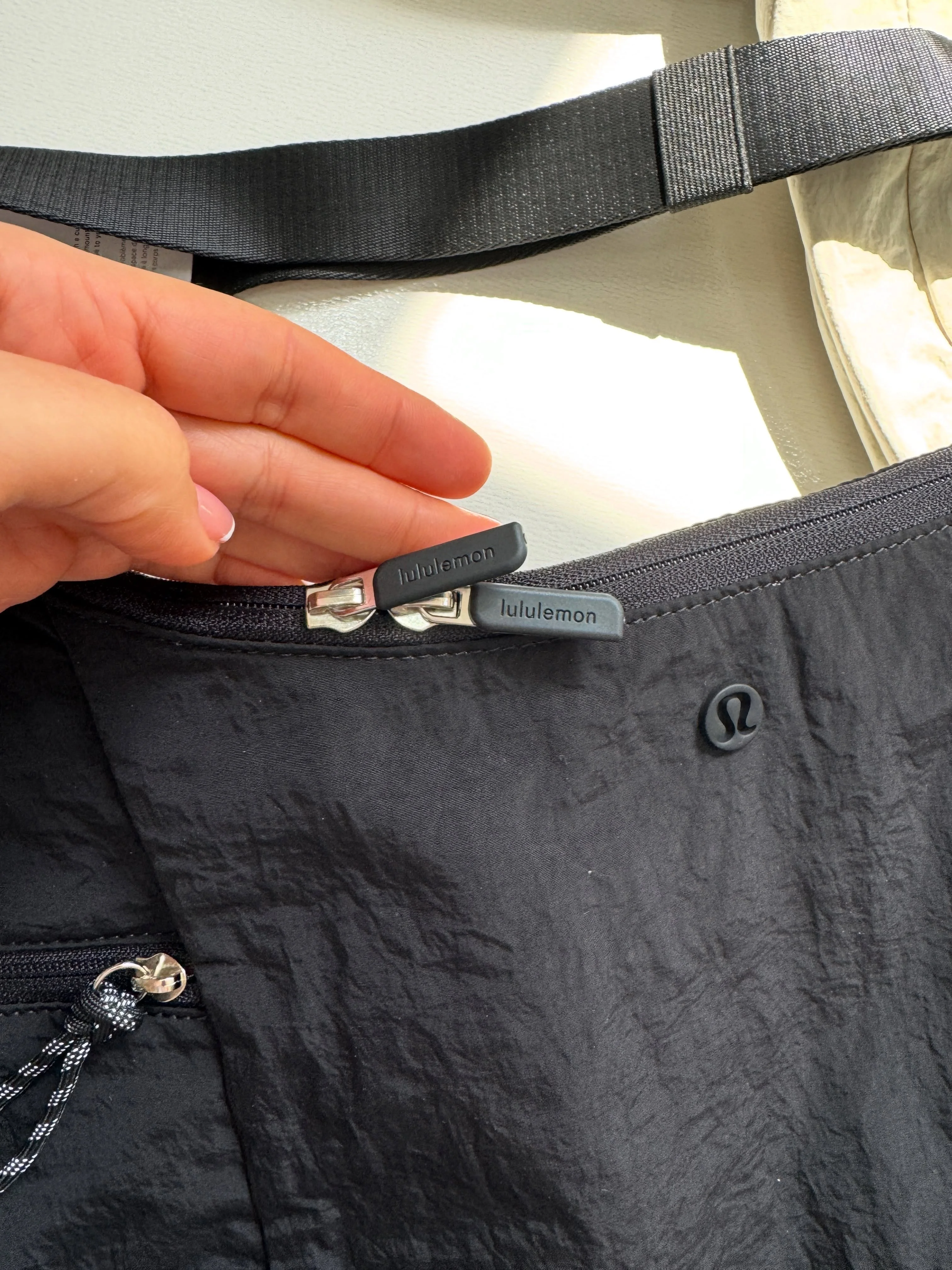 کیف اسلوچی اسلینگ Lululemon 13L مشکی؛ نمای زیپ دهانه، لوگو و قطعه تنظیم بند در دست