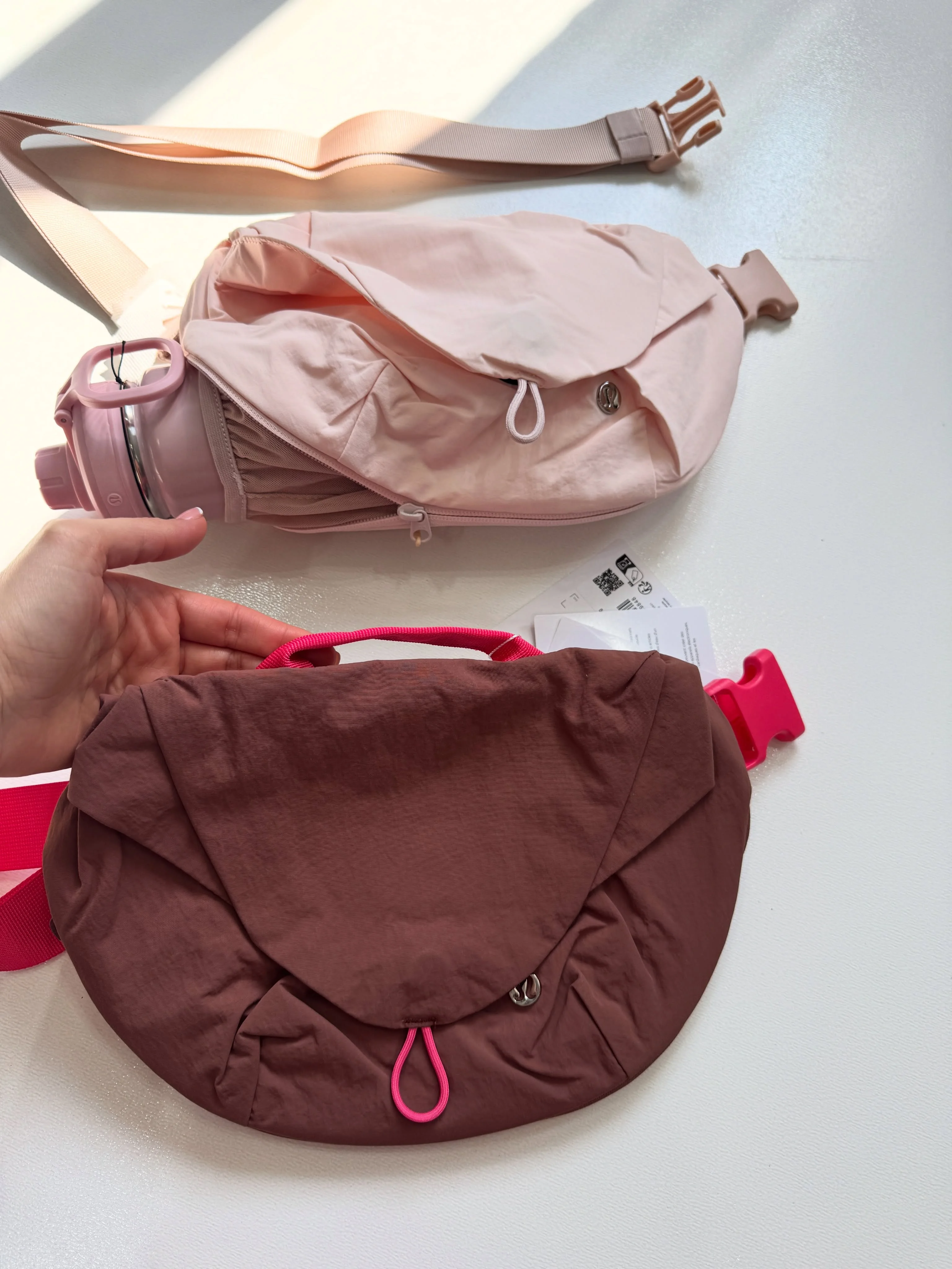 نمای نزدیک جلوی کراس بادی کرو Lululemon قهوه‌ای در دست؛ کیف صورتی با نگهدارنده بطری در پس‌زمینه