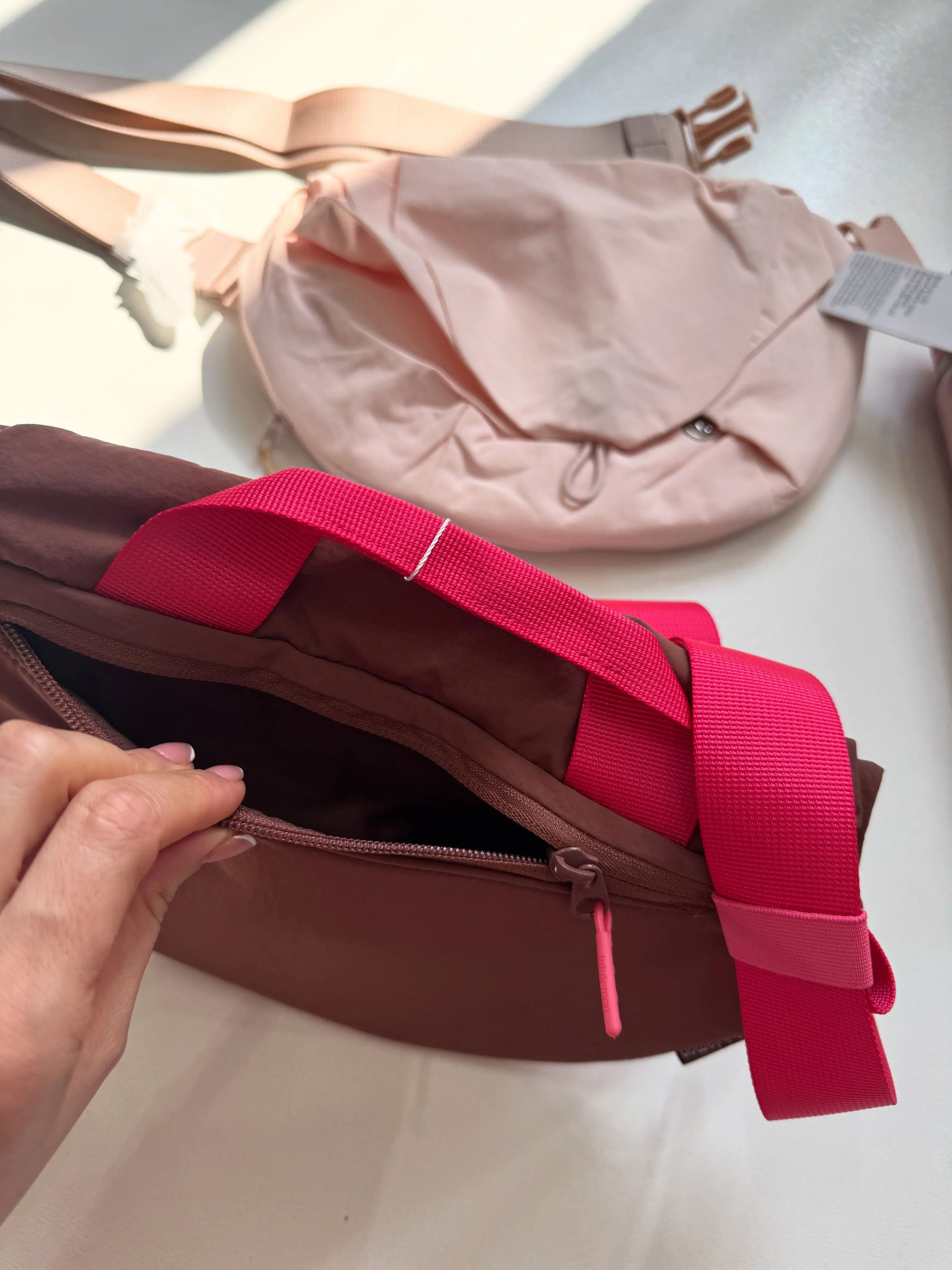 جیب زیپی باز در پشت کراس بادی کرو Lululemon قهوه‌ای با بند سرخابی؛ کیف صورتی در پس‌زمینه