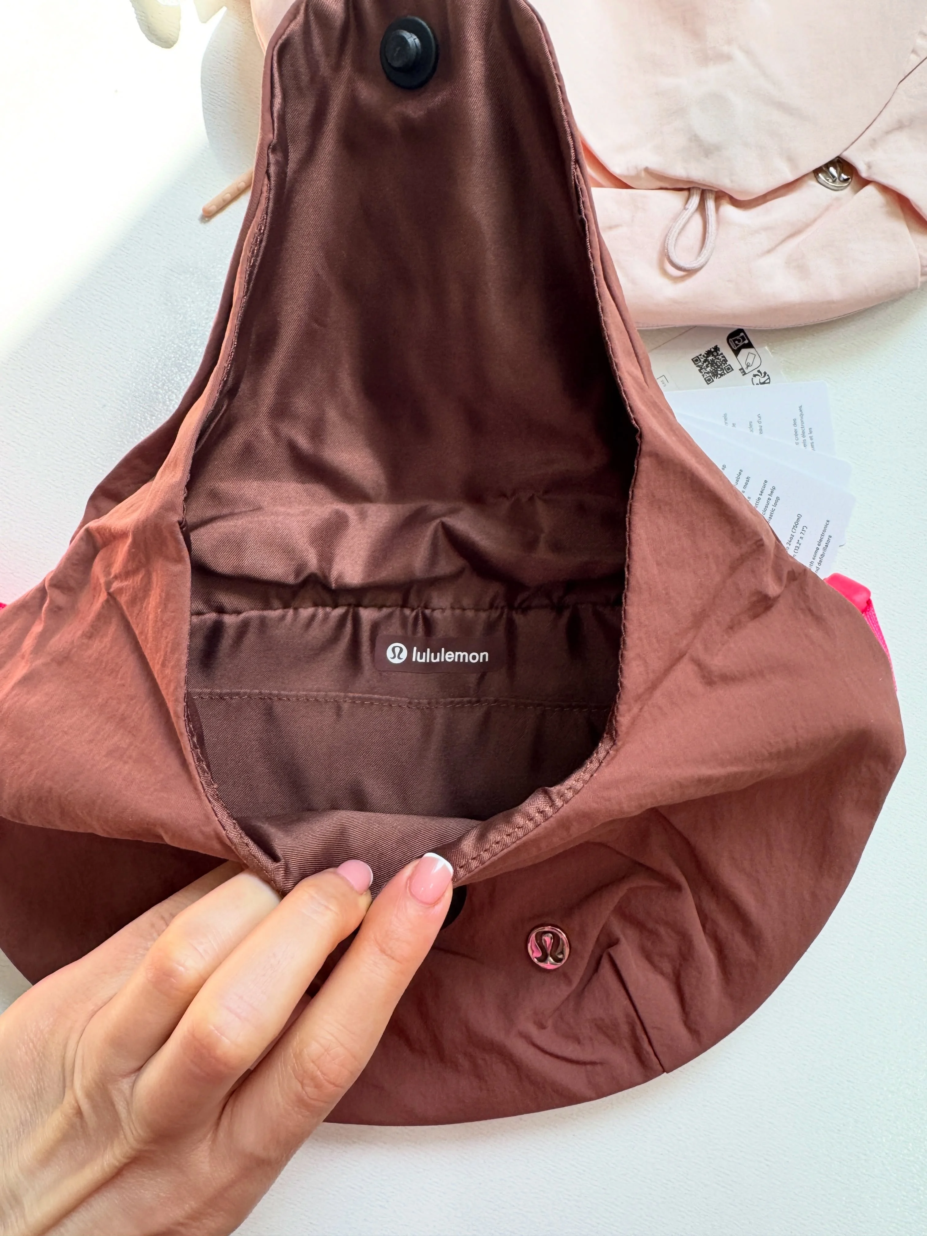 نمای داخل کراس بادی کرو Lululemon قهوه‌ای با جیب زیپی و درپوش باز در دست؛ کیف صورتی در پس‌زمینه