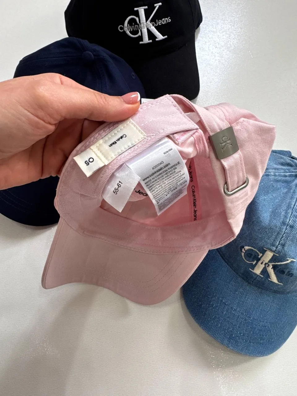 برچسب جنس و سایز کلاه بال کپ Calvin Klein صورتی با نوشته ۱۰۰٪ پنبه، OS و ۵۵ تا ۶۱، کنار سگک فلزی