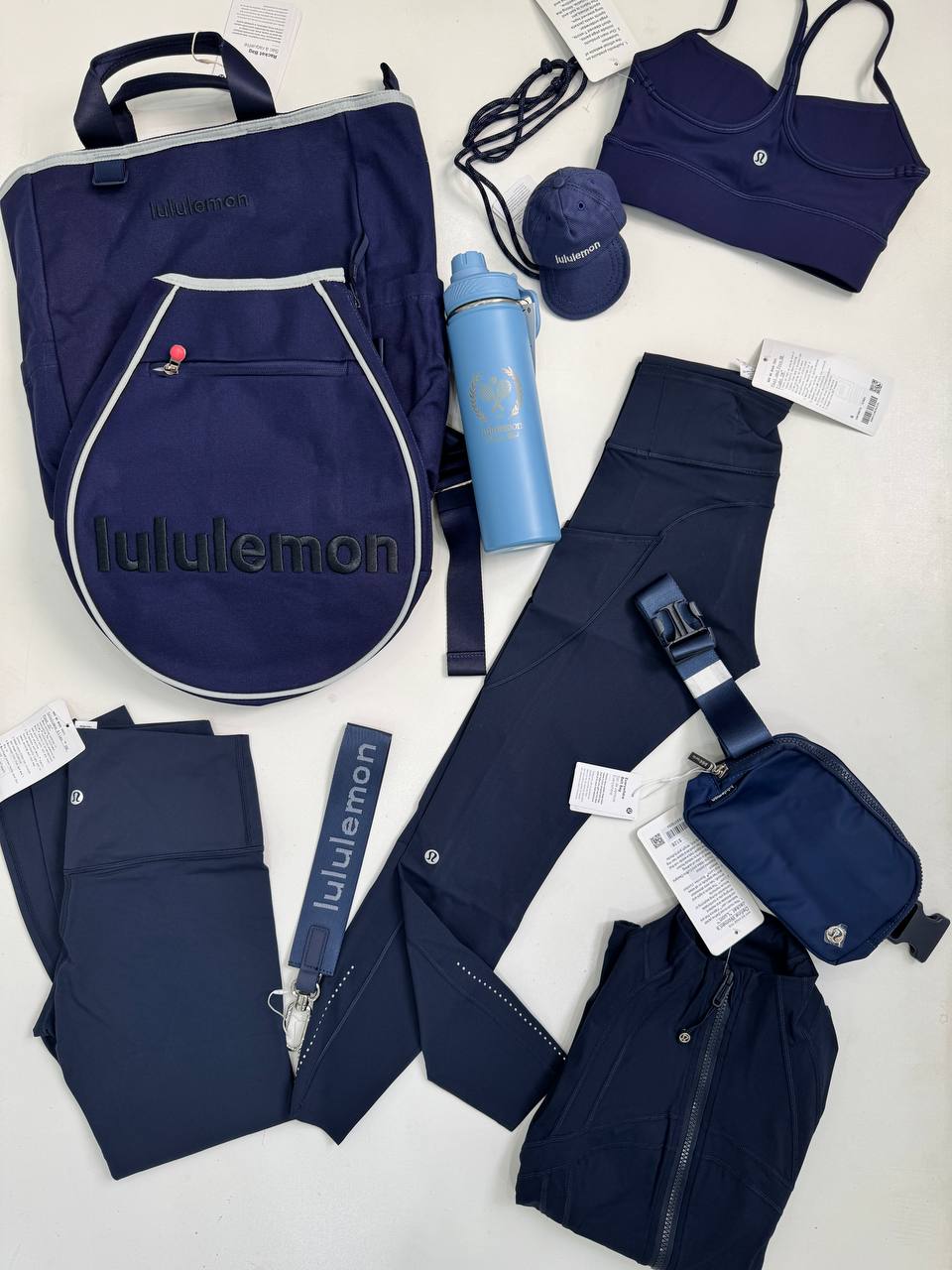 کیف تنیسی راکت بگ Lululemon، کیف تنیس سرمه‌ای و کاور راکت، کنار بطری آبی، نیمتنه، لگ، کیف‌های کوچک و لوازم سرمه‌ای