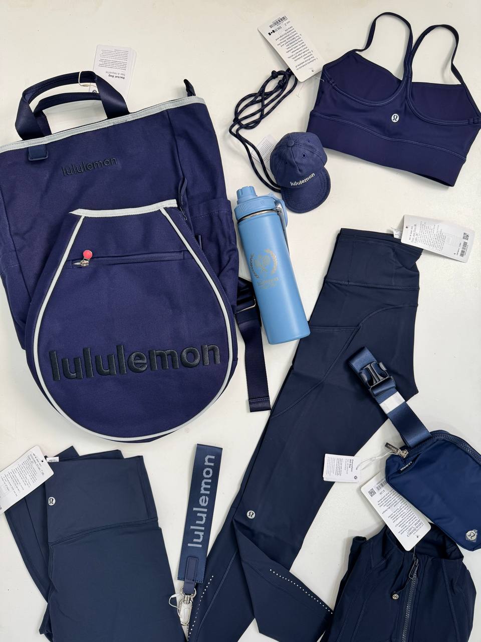 کیف تنیسی راکت بگ Lululemon، کیف تنیس سرمه‌ای و کاور راکت، کنار بطری آبی، نیمتنه، لگ، کیف‌های کوچک و لوازم سرمه‌ای
