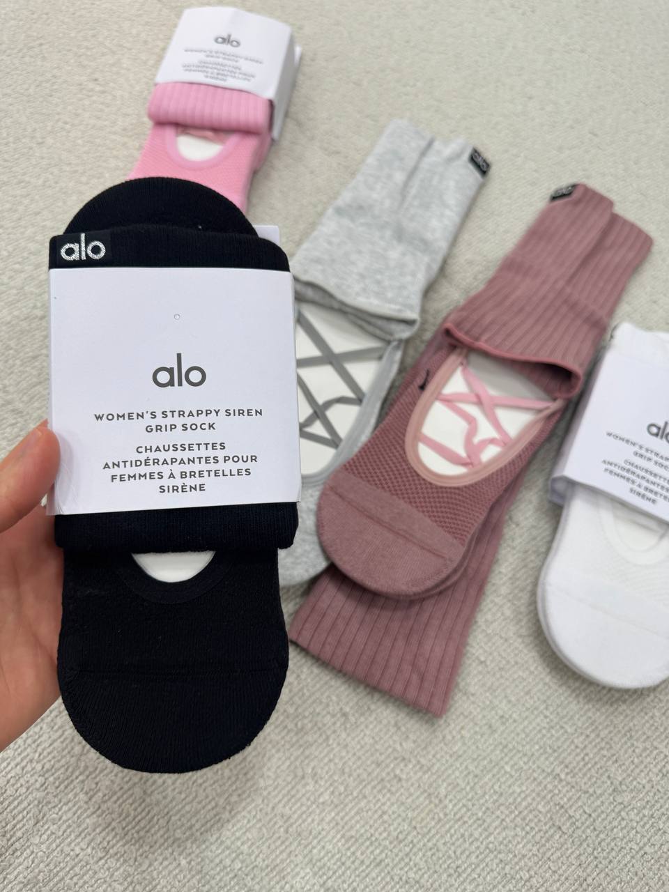 نمای نزدیک بسته‌بندی جوراب پیلاتس ALO مشکی با نوشته Women's Strappy Siren Grip Sock در دست