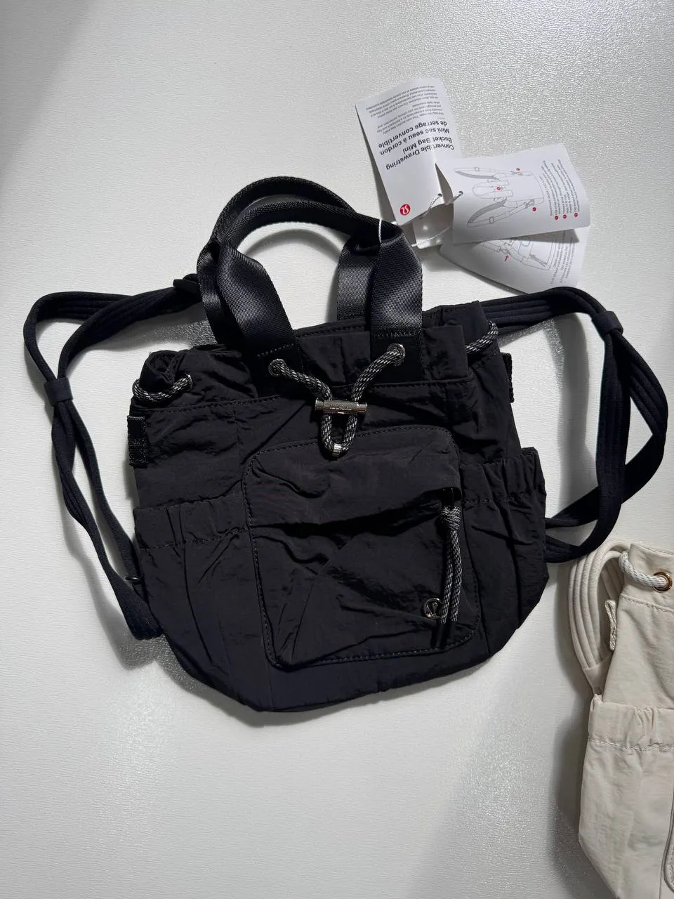 باکت بگ کانورتیبل مینی Lululemon 5L مشکی؛ نمای واقعی جلو و بندهای حمل، با بخشی از کیف شیری کنار آن