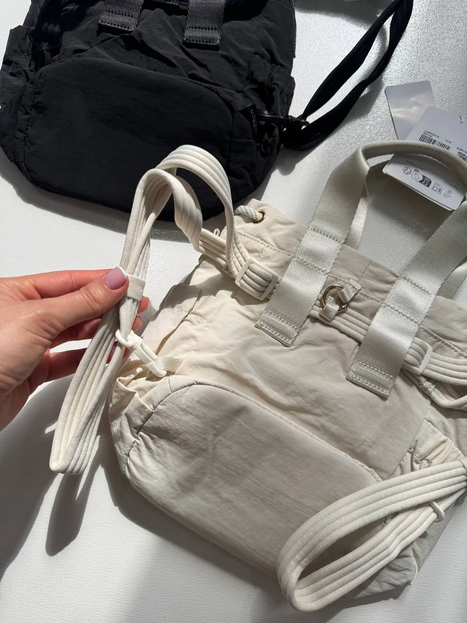 باکت بگ کانورتیبل مینی Lululemon 5L شیری؛ نمای مایل جلو و جیب کناری با بند حمل، در کنار کیف مشکی