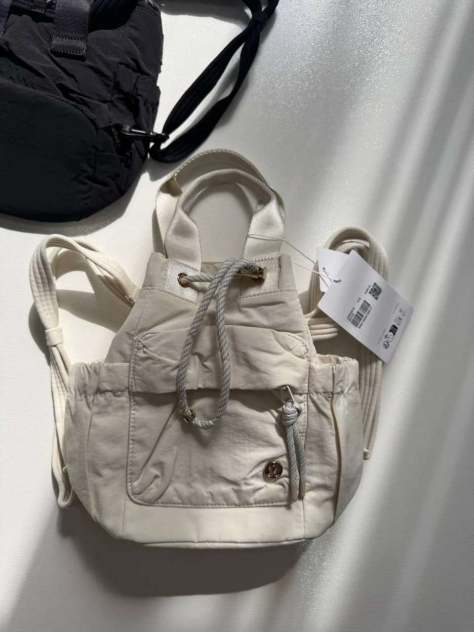 باکت بگ کانورتیبل مینی Lululemon 5L شیری؛ نمای واقعی جلو و بند حمل با بخش کوچکی از کیف مشکی در پس‌زمینه