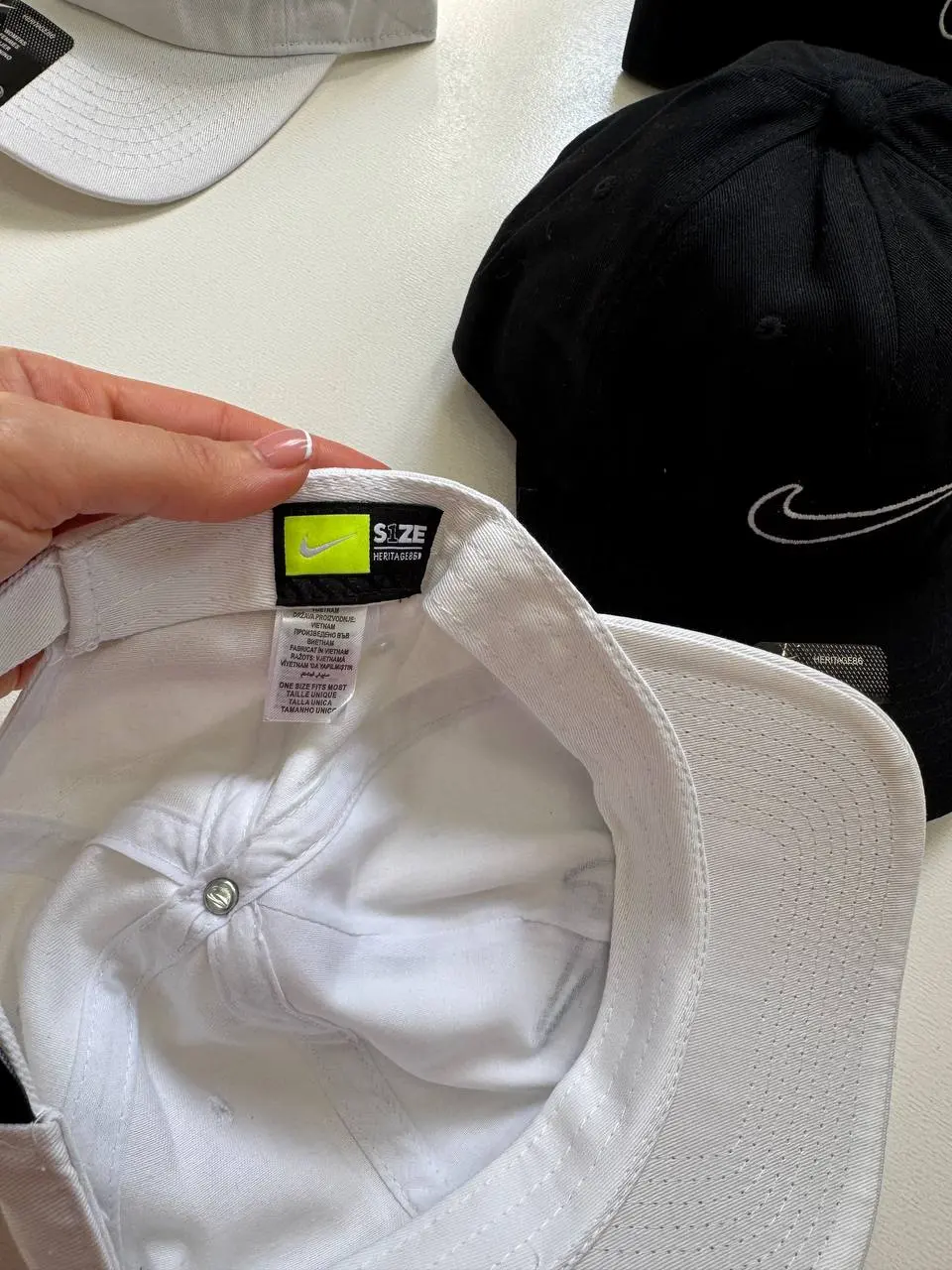 لیبل HERITAGE86 و ONE SIZE FITS MOST داخل کلاه Nike سفید؛ کلاه مشکی در پس‌زمینه
